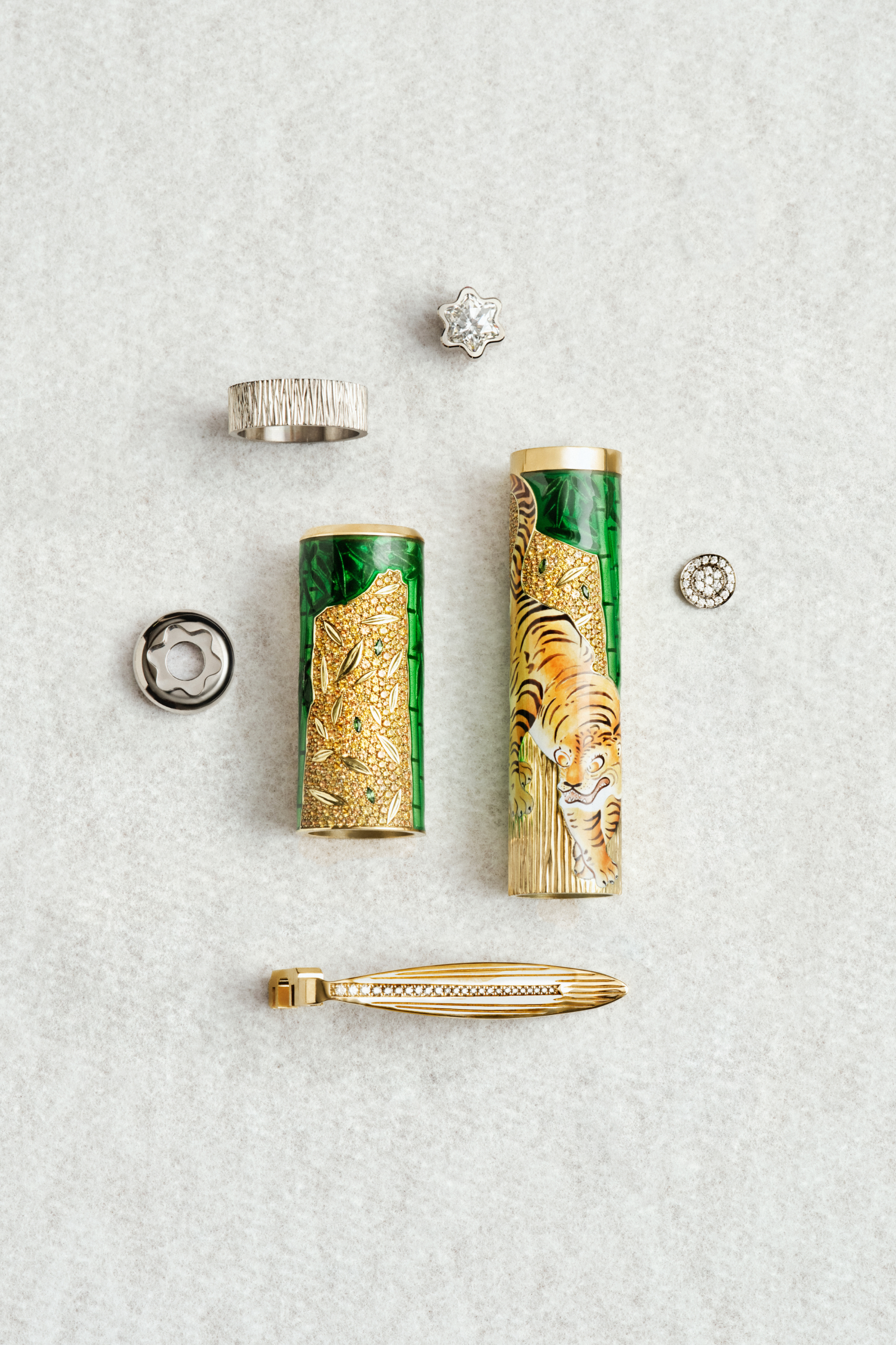Montblanc luxury accessories: gold and green enamel pen barrels, one with tiger motif, a diamond-set clip, and a textured silver ring, on light grey surface.